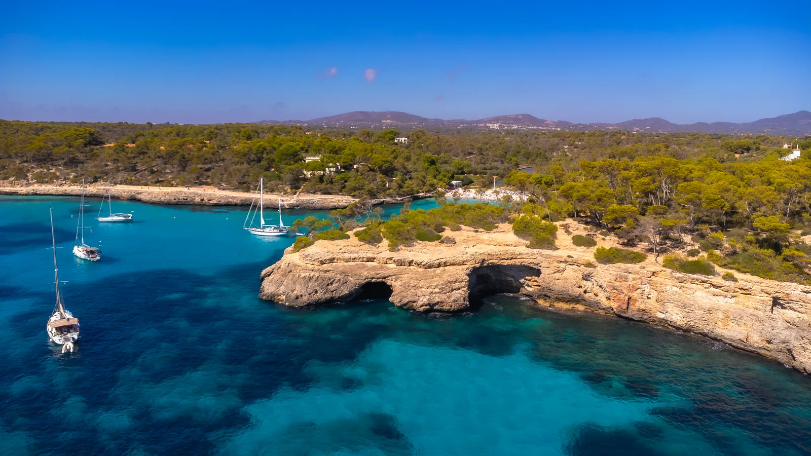 Sailboats anchored in the turquoise water outside Cala Mondrago in Mallorca, with the busy cove and pine covered cliffs behind them.