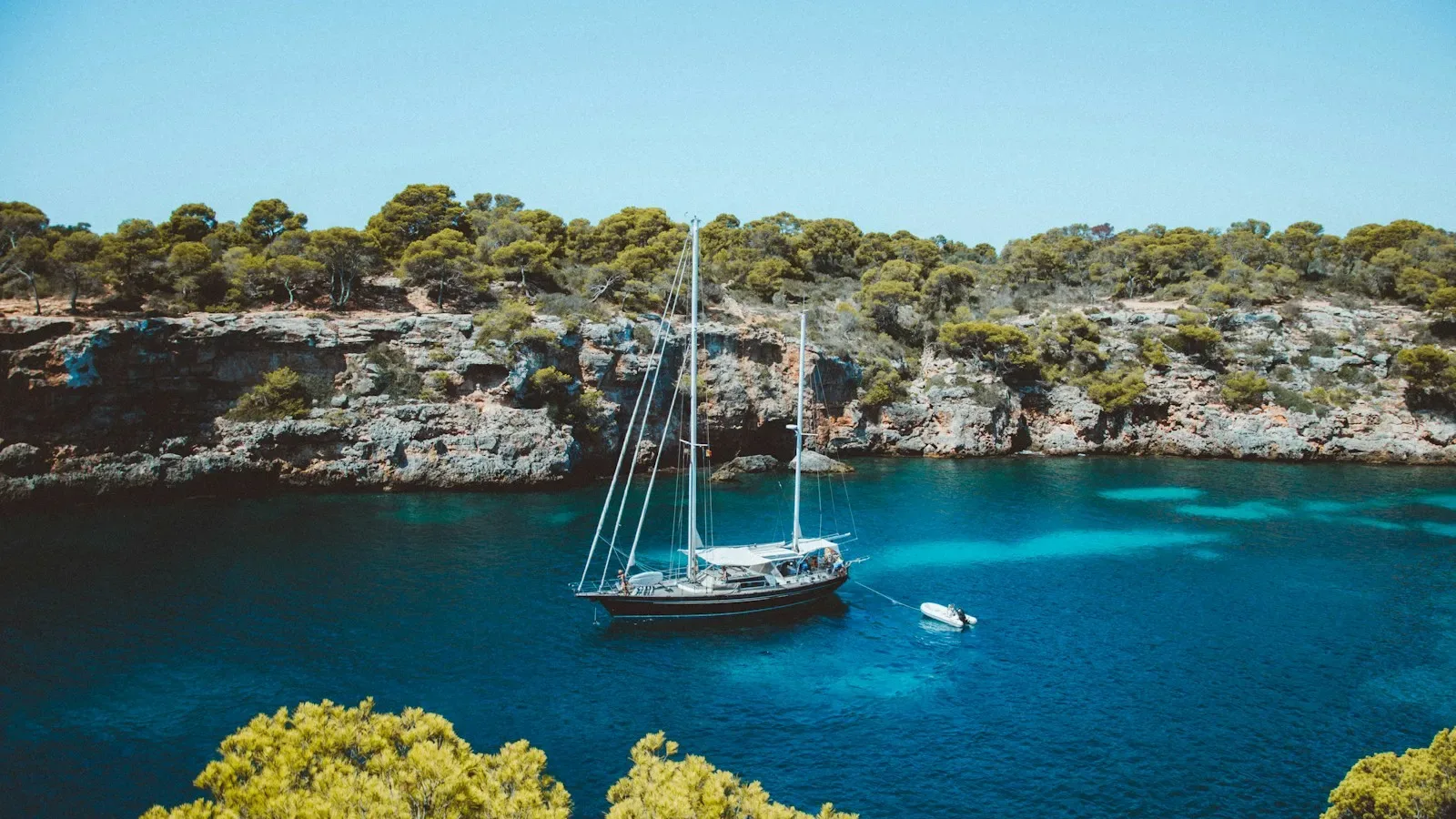 A sailboat anchored in a turquoise cove framed by pine-covered cliffs in the Balearic Islands. Photo by Michael Heuser on Unsplash