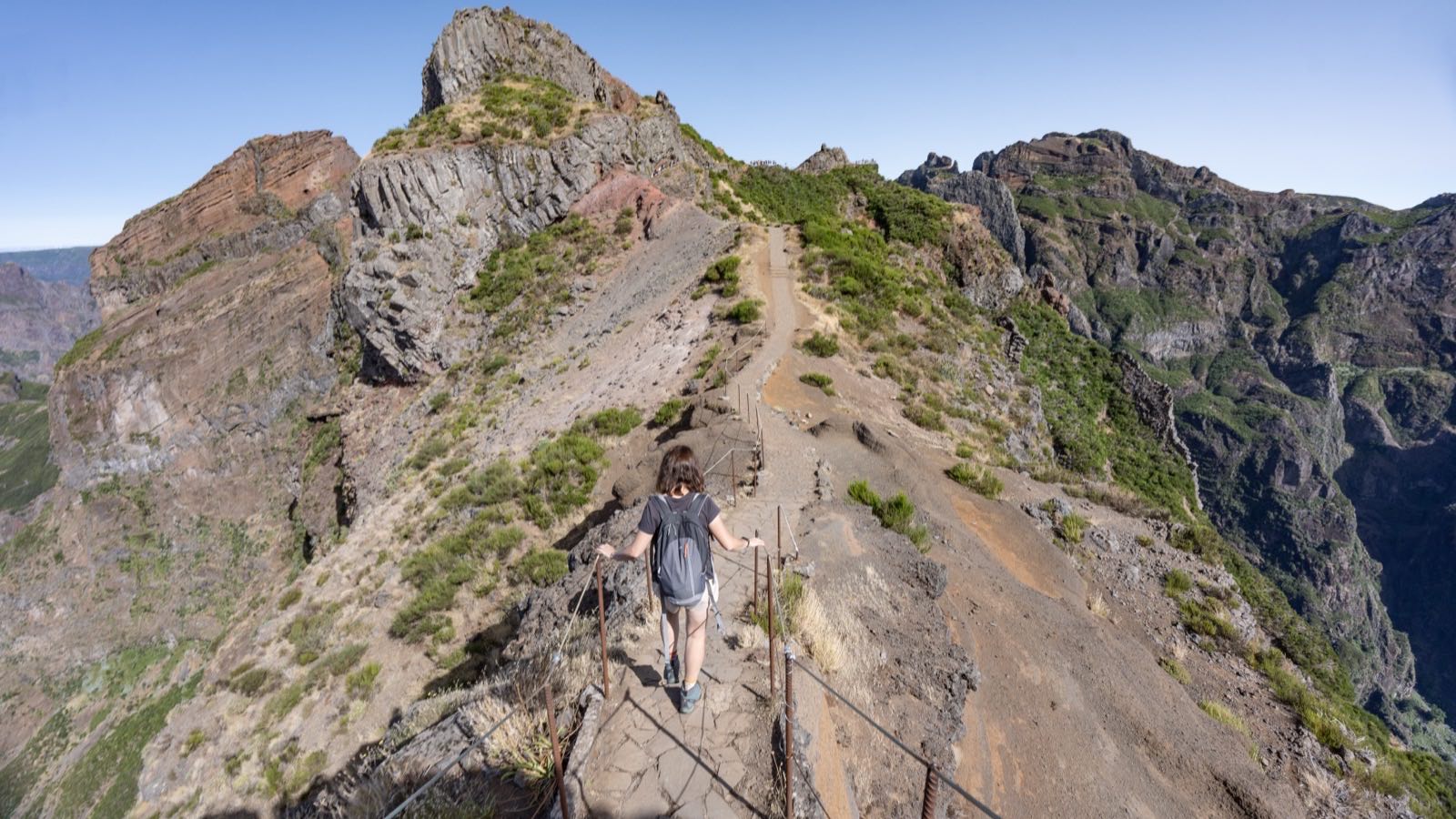 A walker on the narrow rock ridge of the PR1 trail between Pico do Arieiro and Pico Ruivo in Madeira, holding a rope handrail with steep drops on both sides.