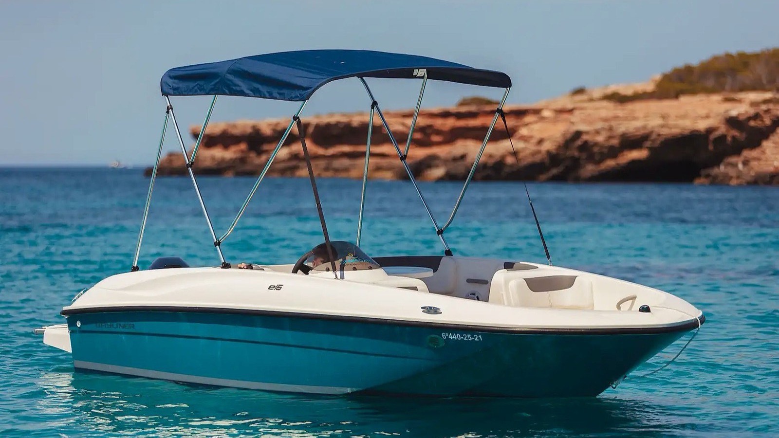 A small white and turquoise licence free rental motorboat with a blue bimini top, anchored in clear water off a low rocky coast in Ibiza.